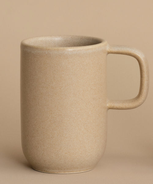 The Pacific Mug, a plain beige stoneware piece with a large handle, rests on a matching surface against a beige background. It features a 10-oz. capacity, blending style and functionality seamlessly.