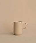 The Pacific Mug, a plain beige stoneware mug with a handle, sits on a matching surface against a beige background. With a 10-oz. capacity, this Pacific Dinnerware piece is perfect for enjoying your favorite hot drinks.