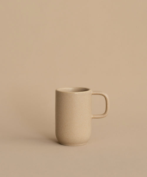 The Pacific Mug, a plain beige stoneware mug with a handle, sits on a matching surface against a beige background. With a 10-oz. capacity, this Pacific Dinnerware piece is perfect for enjoying your favorite hot drinks.