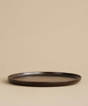 The Pacific Platter, a dark brown round flat stoneware plate with a subtle rim, sits empty on a beige surface against a plain beige background.