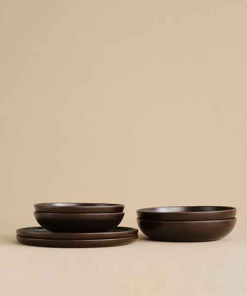 Two dark brown ceramic bowls are stacked on the Pacific Platter, a stoneware serving plate. Set against a plain beige background, this simple and minimalistic arrangement showcases the matte finish of the tableware.