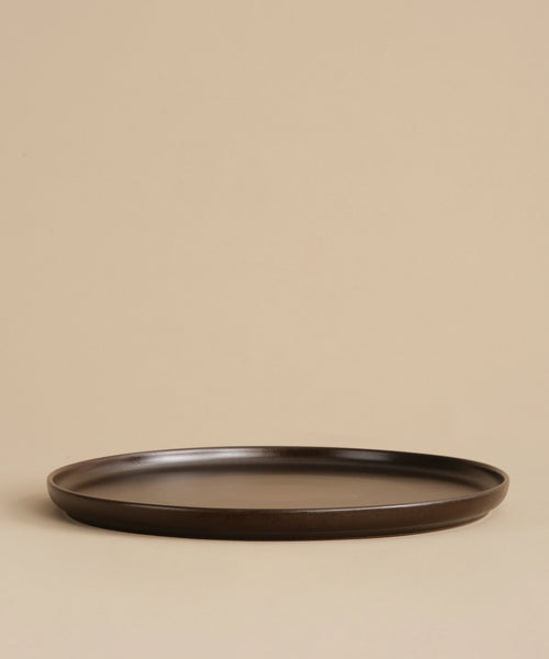The Pacific Platter, a dark brown round flat stoneware plate with a subtle rim, sits empty on a beige surface against a plain beige background.