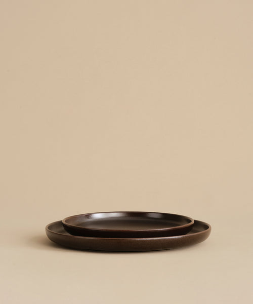 Two nested Pacific Salad Plates in dark brown from the Pacific collection rest on a beige surface against a plain beige background.