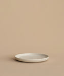 A minimalist, round, white Pacific Salad Plate made of stoneware in Portugal rests empty on a beige surface with a beige background.