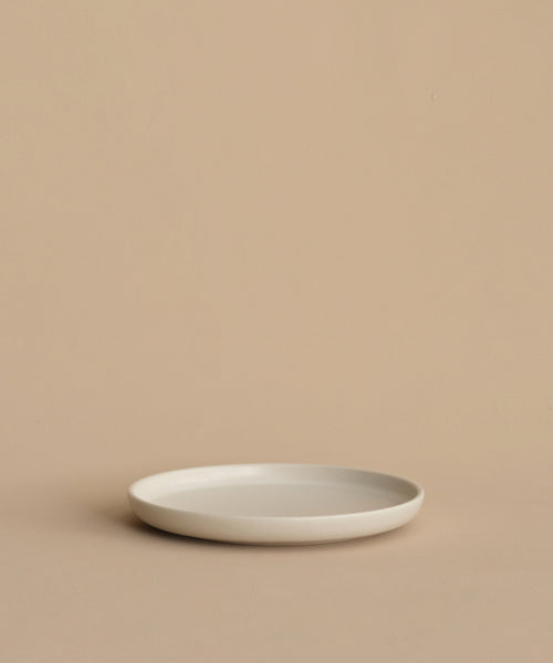 A minimalist, round, white Pacific Salad Plate made of stoneware in Portugal rests empty on a beige surface with a beige background.