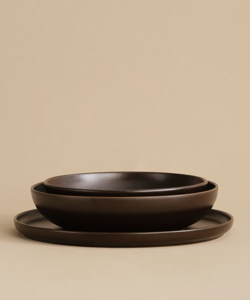 The Pacific Serving Bowl set features three dark brown ceramic pieces of varying sizes—a shallow bowl, a deeper bowl, and a flat plate—stacked together against a beige background. Ideal for adding style to your dinnerware collection.