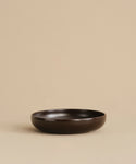 The Pacific Soup Bowl is a shallow, round bowl in dark brown, displayed on a beige surface with a plain beige background.