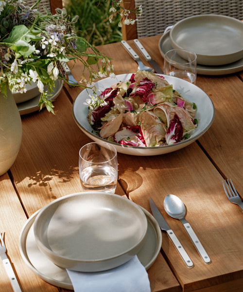 A wooden outdoor table set features Pacific Soup Bowls at the center filled with mixed green and red salad. Glasses and a vase with flowers add charm, while sunlight casts soft shadows over the inviting setting.