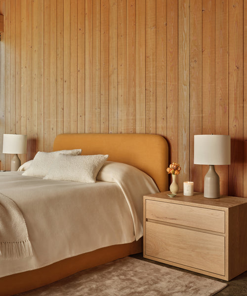 A cozy bedroom with a wooden accent wall, mustard upholstered headboard, cream bedding, the handmade Palms Nightstand in White Oak, two table lamps, a candle, and a small vase of flowers.