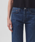 A person wears high-waisted, dark blue Parker Jean by AGOLDE and a black top. The image, cropped from shoulders to upper thighs, shows their relaxed hand at the side against a plain, light background.