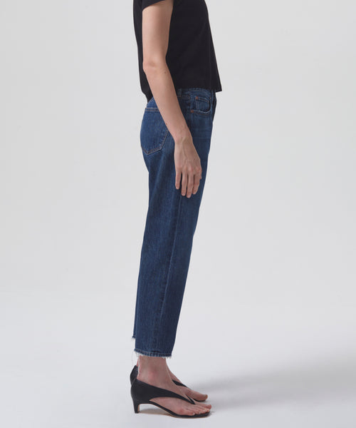 A person stands in profile wearing a black short-sleeve top, Parker Jean vintage high rise denim, and black open-toe wedge sandals against a plain white background.