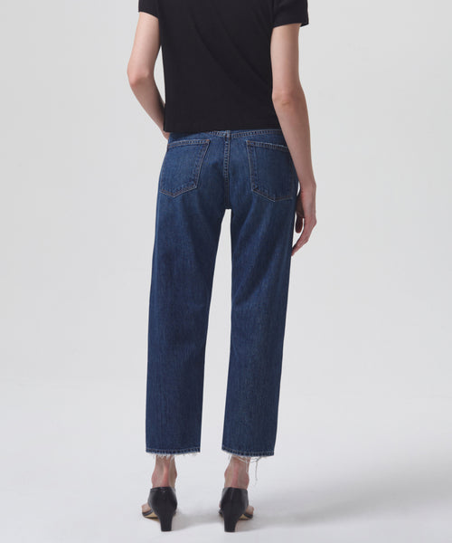 Person standing against a white background, seen from behind, wearing a black short-sleeve shirt, Parker Jean by AGOLDE with raw hems in a high-rise fit, and black heeled sandals.