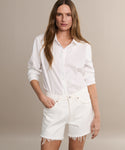 A woman with long brown hair wears a white button-up shirt with rolled sleeves and Parker Long Short denim shorts, standing with one hand in her pocket against a plain beige background.