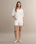 A woman stands against a neutral background wearing a white button-up shirt with rolled sleeves and AGOLDE Parker Long Shorts featuring a frayed hem. She completes the look with beige flat shoes, relaxed posture, and one hand in her pocket.