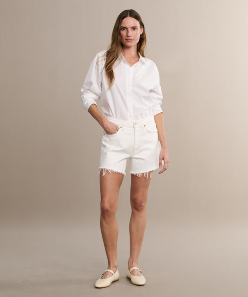 A woman stands against a neutral background wearing a white button-up shirt with rolled sleeves and AGOLDE Parker Long Shorts featuring a frayed hem. She completes the look with beige flat shoes, relaxed posture, and one hand in her pocket.