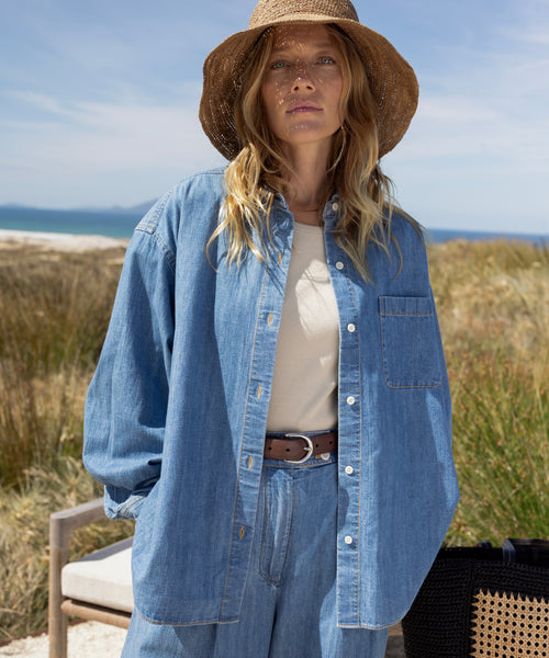 A woman wearing the Parker Oversized Shirt stands outdoors with grass, sand dunes, and blue sky in the background. She pairs the summer-weight denim shirt with a wide-brimmed straw hat, her loose wavy hair framing her neutral expression.