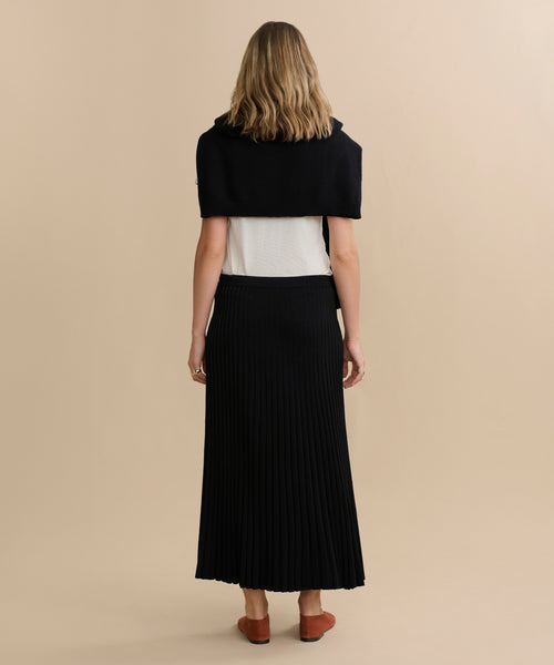 A woman stands with her back to the camera, wearing a white top, black sweater over her shoulders, long black Penelope Skirt, and brown flats against a plain beige background.