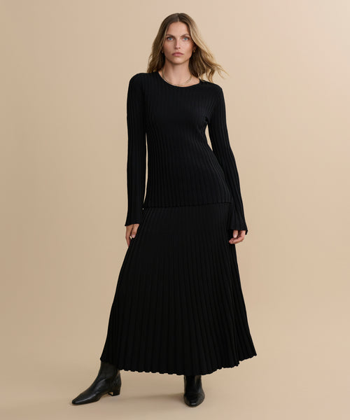 A woman stands against a beige background wearing a black ribbed-knit long-sleeve top paired with the Penelope Skirt, an ankle-length rib skirt, and black ankle boots. She faces forward with one hand on her hip and her hair down.