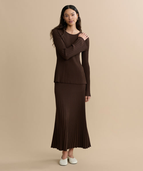 A woman with long dark hair stands against a beige background, wearing the Penelope Skirt—a long, ribbed brown knit skirt—with a matching brown ribbed top and white slip-on shoes, her expression neutral.