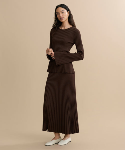 A woman with long dark hair stands against a beige background wearing a ribbed brown top with flared cuffs and the Penelope Skirt—a matching long, pleated skirt—paired with classic white shoes.