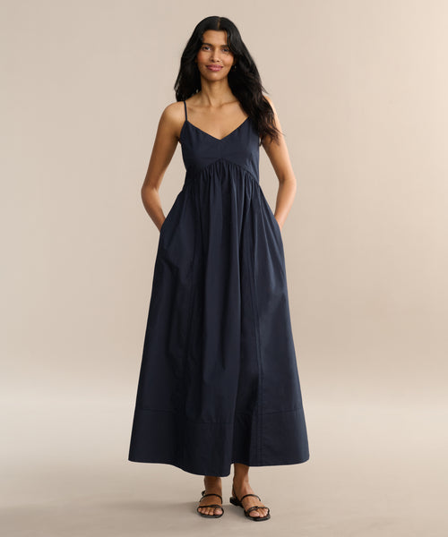 A woman with long, wavy dark hair smiles softly as she stands against a plain light background, wearing the sleeveless Peninsula Dress with thin straps and pockets, paired with sandals. Her hands rest in the dress’s pockets.