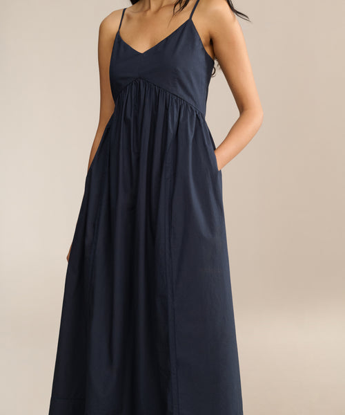 A woman stands against a beige background, wearing the Peninsula Dress—a sleeveless navy sundress with thin straps and pockets.