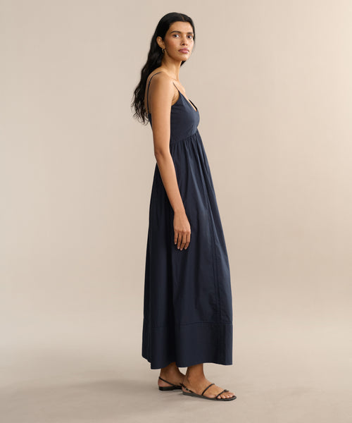 A woman with long dark hair stands sideways wearing the sleeveless Peninsula Dress in navy Italian washed poplin and black sandals, set against a plain beige background.