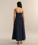 A woman with long dark hair stands facing away, wearing the sleeveless Peninsula Dress in navy Italian washed poplin with thin straps and flat sandals. The background is plain and neutral.
