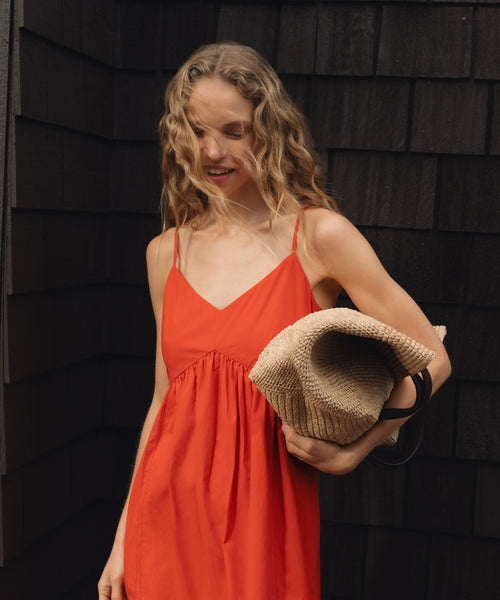 Woman in an red dress holding a beige woven raffia handbag against a dark wooden wall.