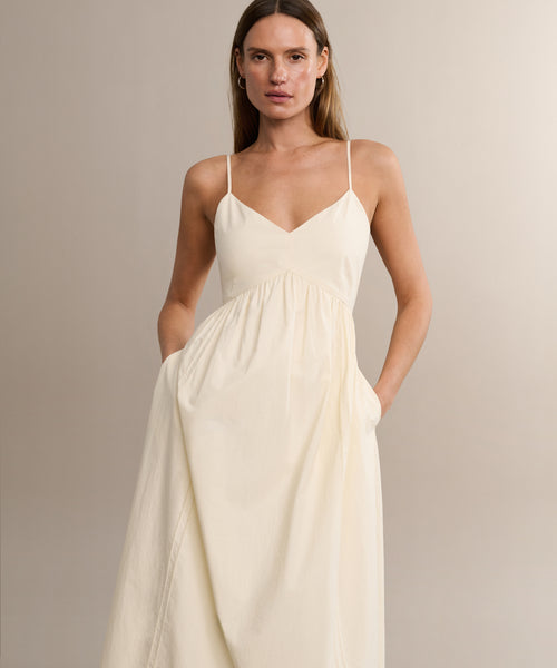 A woman stands against a neutral background, hands in her pockets, wearing the Peninsula Dress—a sleeveless, cream maxi sundress made from Italian washed poplin with thin straps.