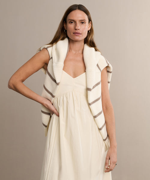 A woman with long brown hair wears the sleeveless Peninsula Dress, with a cream and brown striped knit sweater draped over her shoulders, set against a plain beige background.