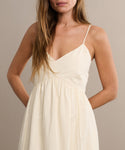 A woman stands against a beige background with her hands in the pockets of the Peninsula Dress, a sleeveless sundress with thin spaghetti straps. Her face is partially out of frame.