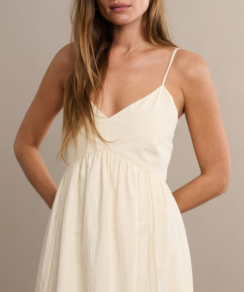 A woman stands against a beige background with her hands in the pockets of the Peninsula Dress, a sleeveless sundress with thin spaghetti straps. Her face is partially out of frame.