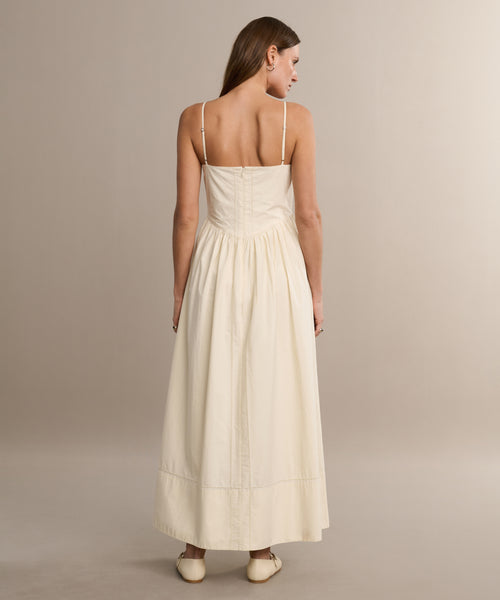A woman with straight brown hair faces away, wearing the Peninsula Dress—a sleeveless, cream sundress in Italian washed poplin with thin straps and a gathered waist—paired with cream shoes, set against a neutral background.