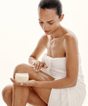 A woman in a white towel applies Perfecting Body Scrub from Oak Essentials to her leg, sitting against a light background and focusing on her hydrating skincare routine.