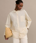 A woman in the Pierce Shirt, styled with white pants and a woven straw bag, stands against a beige background, looking down with her hair pulled back.