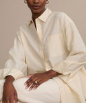 Seated against a beige backdrop, a person wears the loose Pierce Shirt crafted from textured Italian cotton, paired with matching pants. Gold hoop earrings and rings accent their look, as their hand rests on their knee and their face is partially visible.
