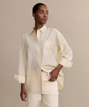 A woman with short, slicked-back hair and small hoop earrings poses confidently in a cream Pierce Shirt made of textured cotton, sleeves rolled up, paired with light pants, set against a plain beige background.