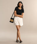 A woman smiles with arms crossed, wearing a black t-shirt, Pleated Short in white, black sandals, and a black woven shoulder bag against a neutral background.
