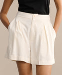 A person wearing Pleated Short in ivory raw silk, high-waisted with side pockets, and a black sleeveless top stands with hands in the shorts’ pockets against a neutral background.