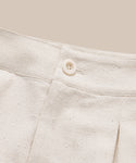 Close-up of the waistband and button closure of the Pleated Short in cream-colored raw silk with a subtle dotted pattern, set against a matching beige background.