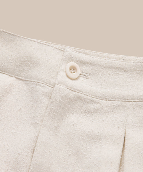 Close-up of the waistband and button closure of the Pleated Short in cream-colored raw silk with a subtle dotted pattern, set against a matching beige background.