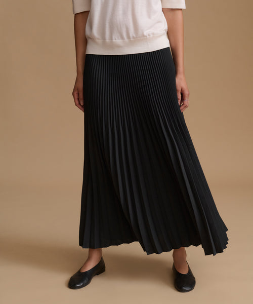 A model wearing a long black pleated skirt, paired with a cream short-sleeve top and black leather flats, set against a solid beige studio background.