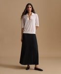 A woman stands against a warm beige studio background wearing a cream short-sleeve collared knit top and a long black pleated skirt, paired with black flats.