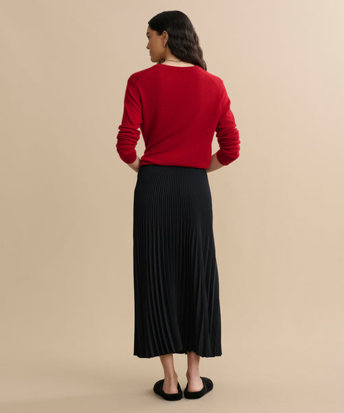 A woman stands with her back to the camera, wearing a black Pleated Skirt, red long-sleeve top, and black shoes. Her loose, wavy dark hair contrasts against a plain beige background.
