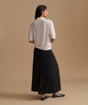 A woman is shown from the back wearing a cream short-sleeve knit top and a long black pleated skirt, standing against a solid tan studio background.