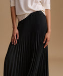 A close-up of a person wearing a black pleated skirt with a cream-colored top, standing against a solid beige background.