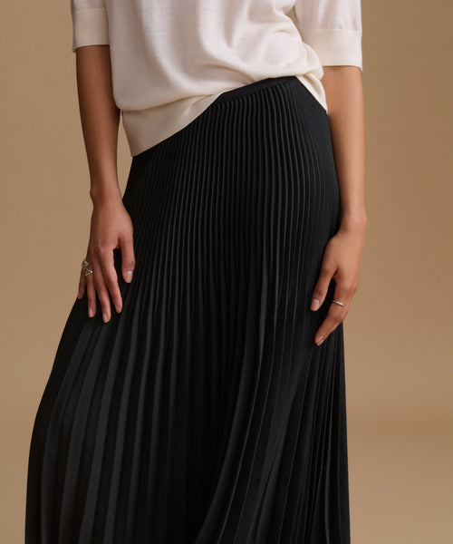 A close-up of a person wearing a black pleated skirt with a cream-colored top, standing against a solid beige background.