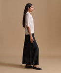 A woman in side profile wearing a white short-sleeve blouse and a black pleated skirt, standing against a solid beige studio background.