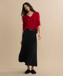 Against a plain beige background, a woman with long dark hair poses casually in a black Pleated Skirt with a belt, a red v-neck sweater, black shoes, and layered necklaces.